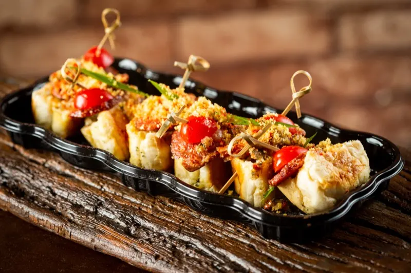 Bruschettas de choripán com tomate-cereja em travessa preta sobre madeira.
