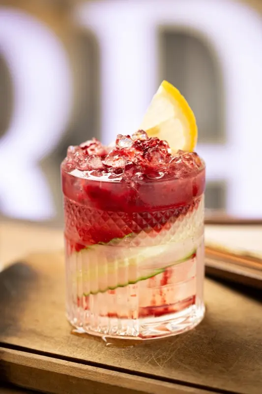 Drink Bramble em copo baixo, com gelo picado, amora e uma fatia de limão.