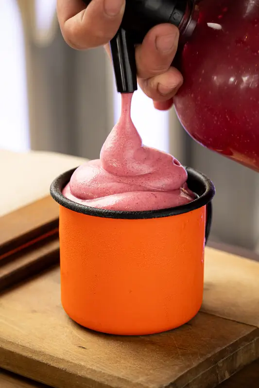 Mão finalizando um drink numa caneca de metal esmaltada laranja, com espuma rosa saindo de um sifão.