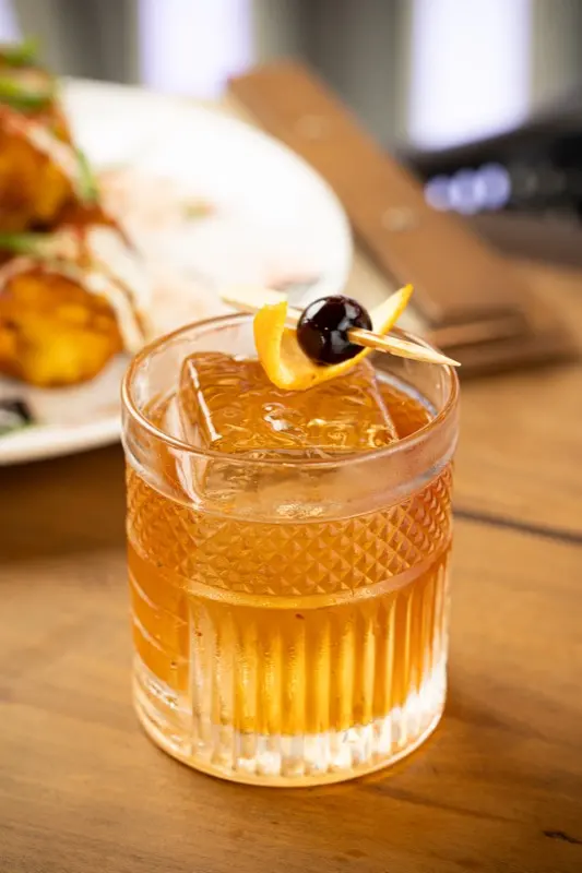 Old Fashioned em copo de vidro lapidado, com uma pedra de gelo, casca de laranja e uma cereja no palito.