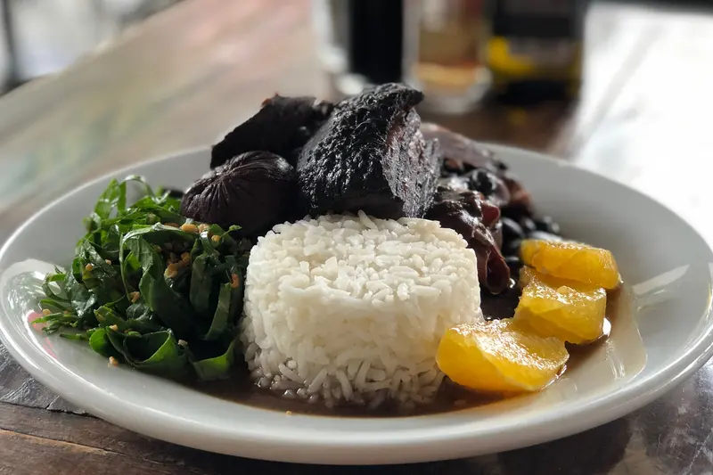 Prato de feijoada com arroz, couve refogada, gomos de laranja e caldo escuro com feijão-preto.